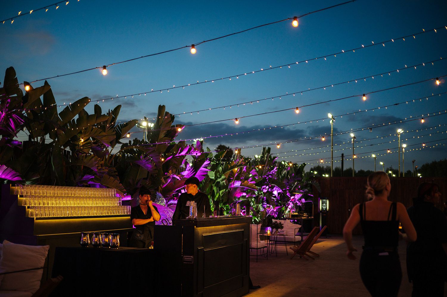 Outdoor evening event venue with string lights, tropical plants, and purple floral arrangements.