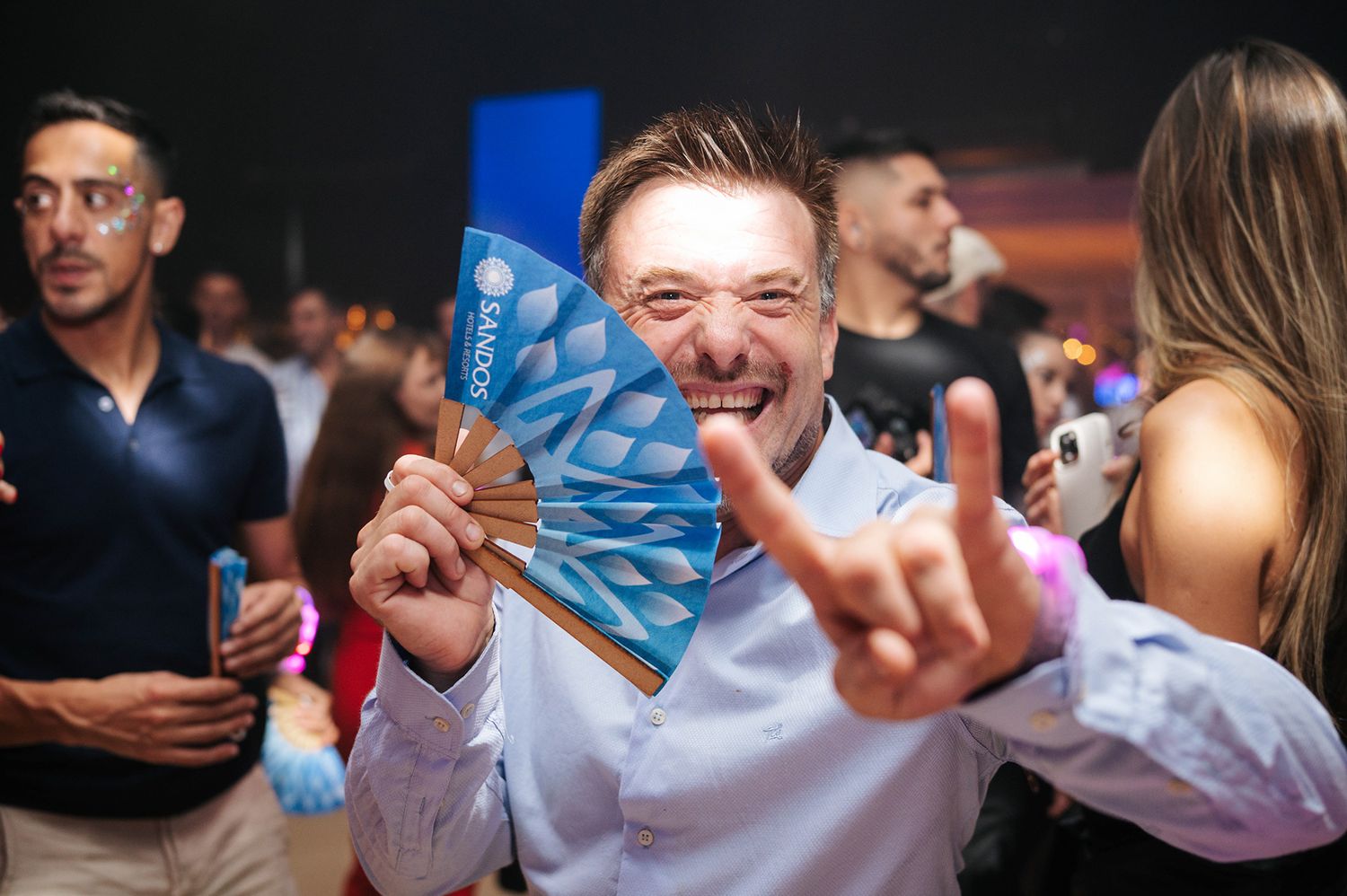 Excited man holding a decorative fan and making rock sign at a vibrant nightclub party event.