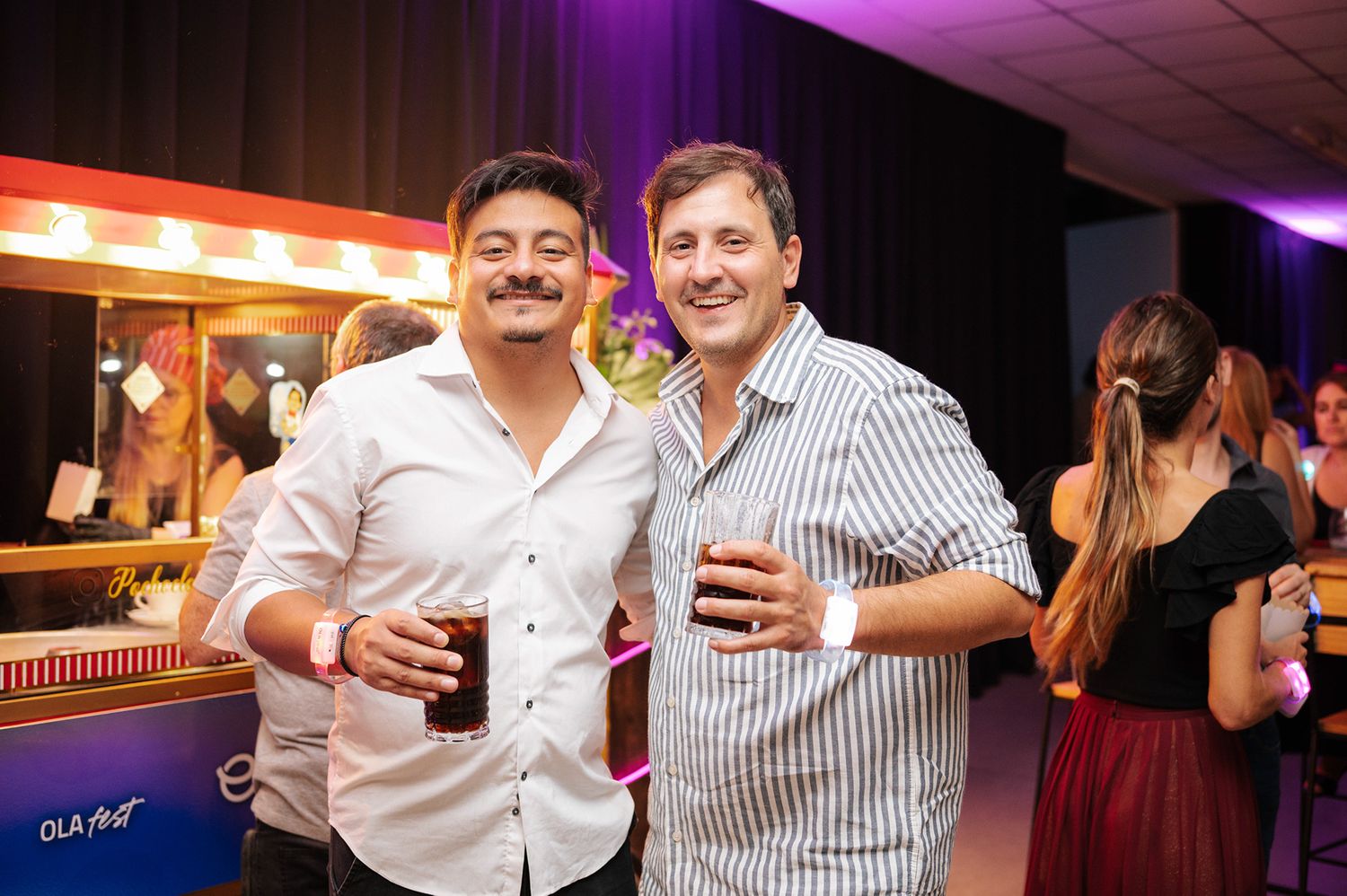 Two smiling men holding drinks together at a colorful indoor party or nightclub event.
