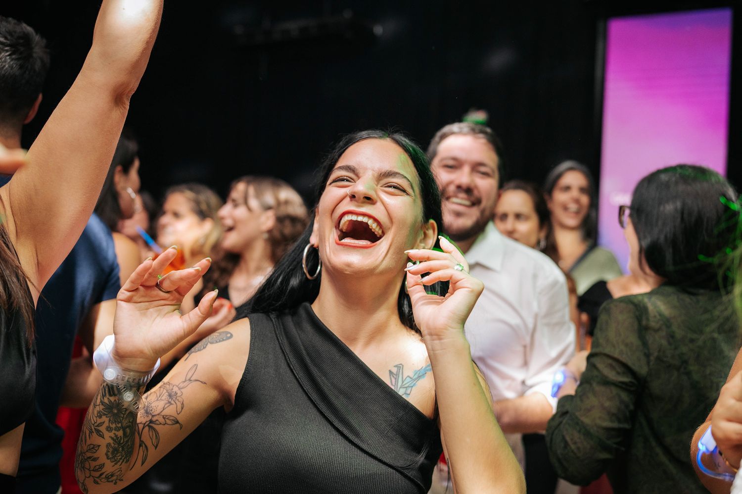 Woman laughing joyfully with tattoos visible at a lively indoor celebration or concert event.