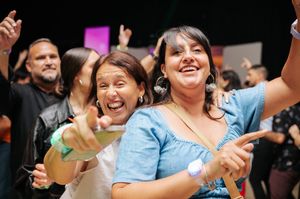 Two women laughing and pointing at the camera while dancing at a lively indoor event.