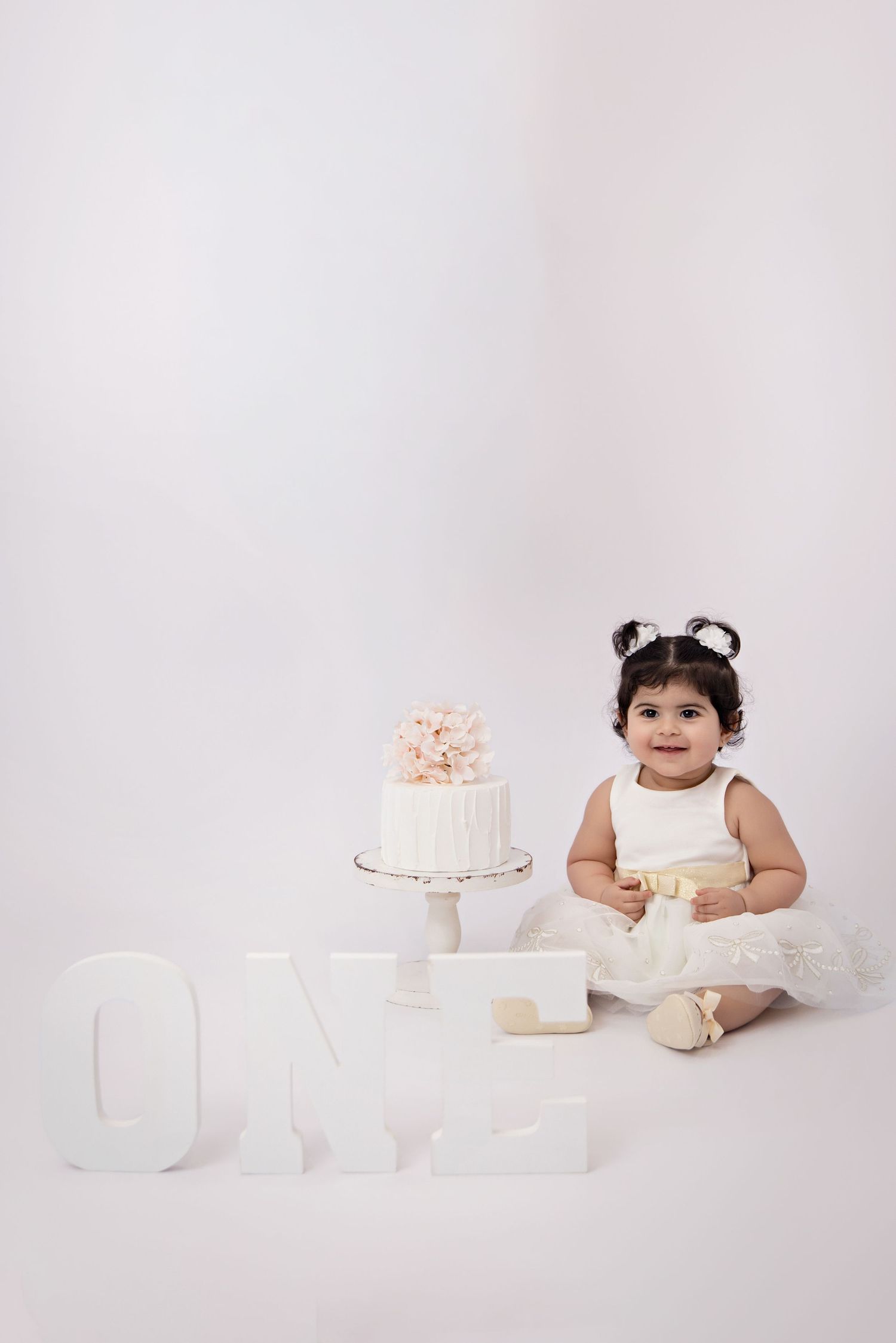 Baby girl in white dress sits beside first birthday cake with ONE letters in bright studio photo.