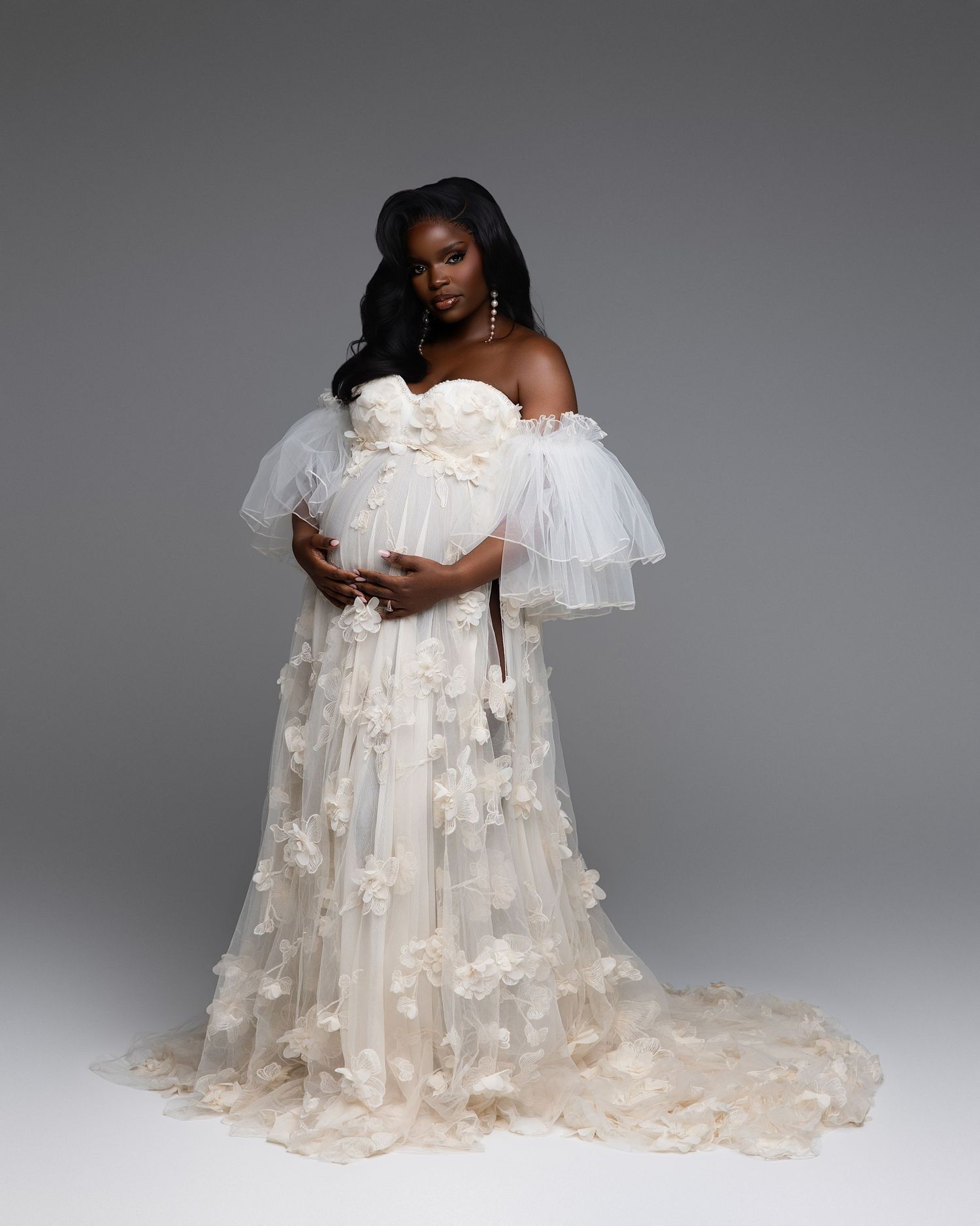 Pregnant woman in elegant white off-shoulder tulle gown with floral appliques, posing gracefully in a studio setting.