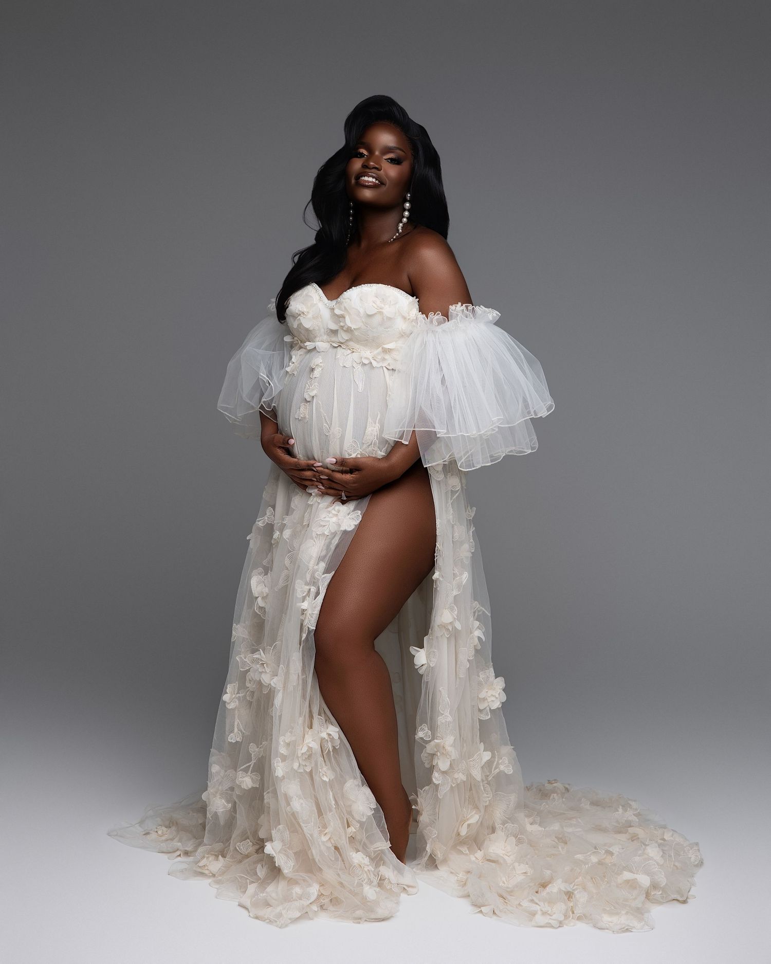 Pregnant woman in white floral tulle maternity gown posing gracefully in a professional studio photo shoot.