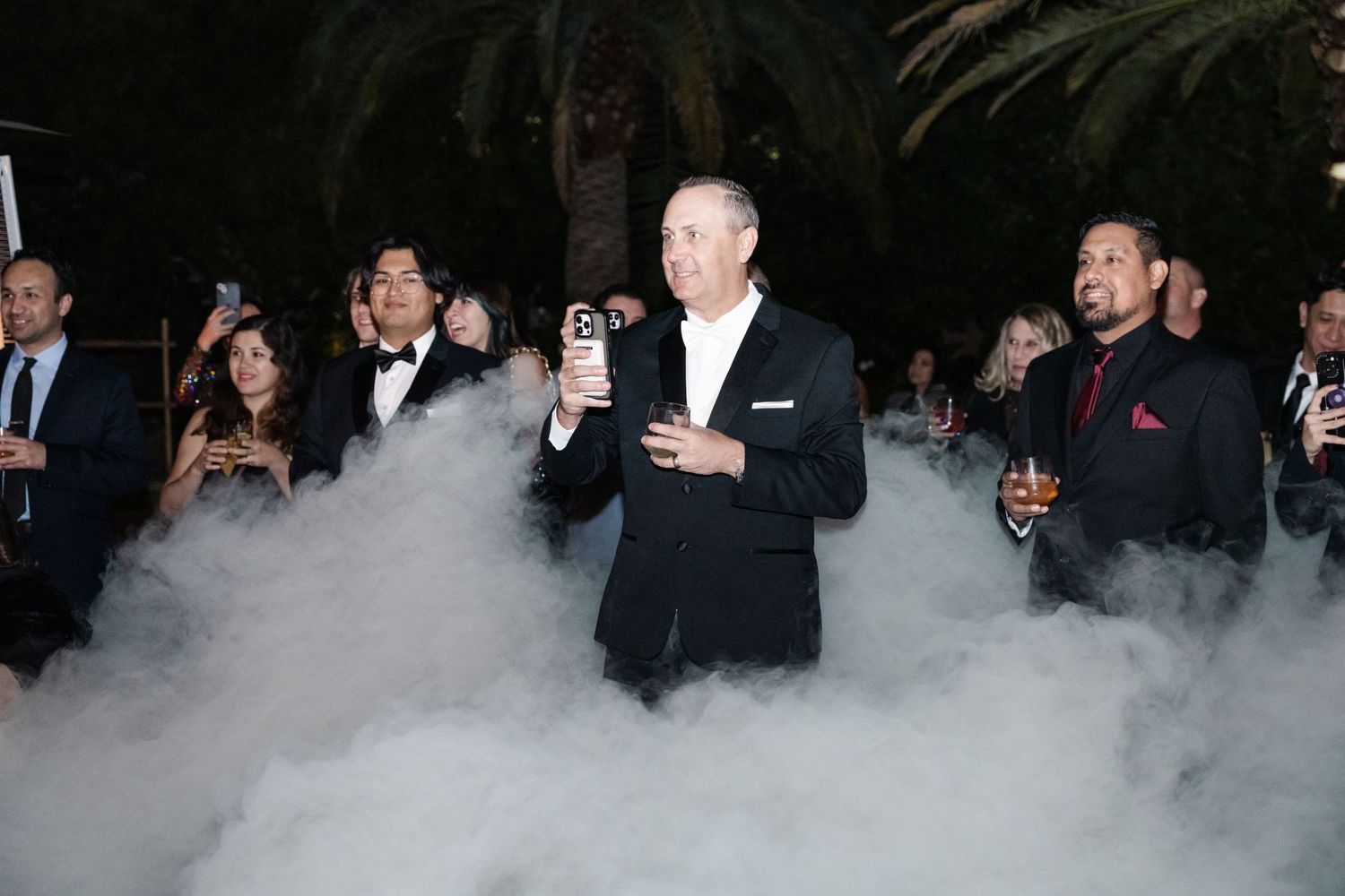 Man in tuxedo holds drink amid dramatic dry ice fog at elegant outdoor evening gala event.