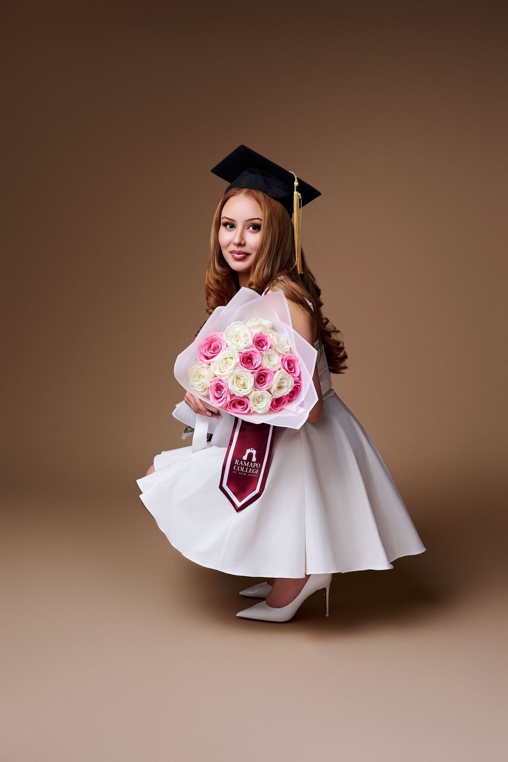 Studio graduation portrait of a Ramapo College RCNJ graduate crouching with a flower bouquet, captured by a North NJ portrait photographer.