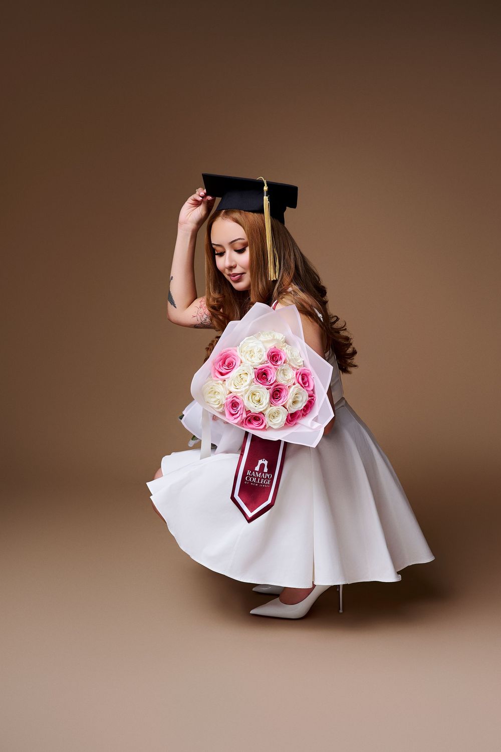 Studio graduation portrait of an RCNJ graduate crouching with a flower bouquet and adjusting her cap, by a North NJ portrait photographer.