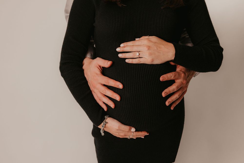 Couple placing hands on pregnant belly wearing a black ribbed dress against a light background.
