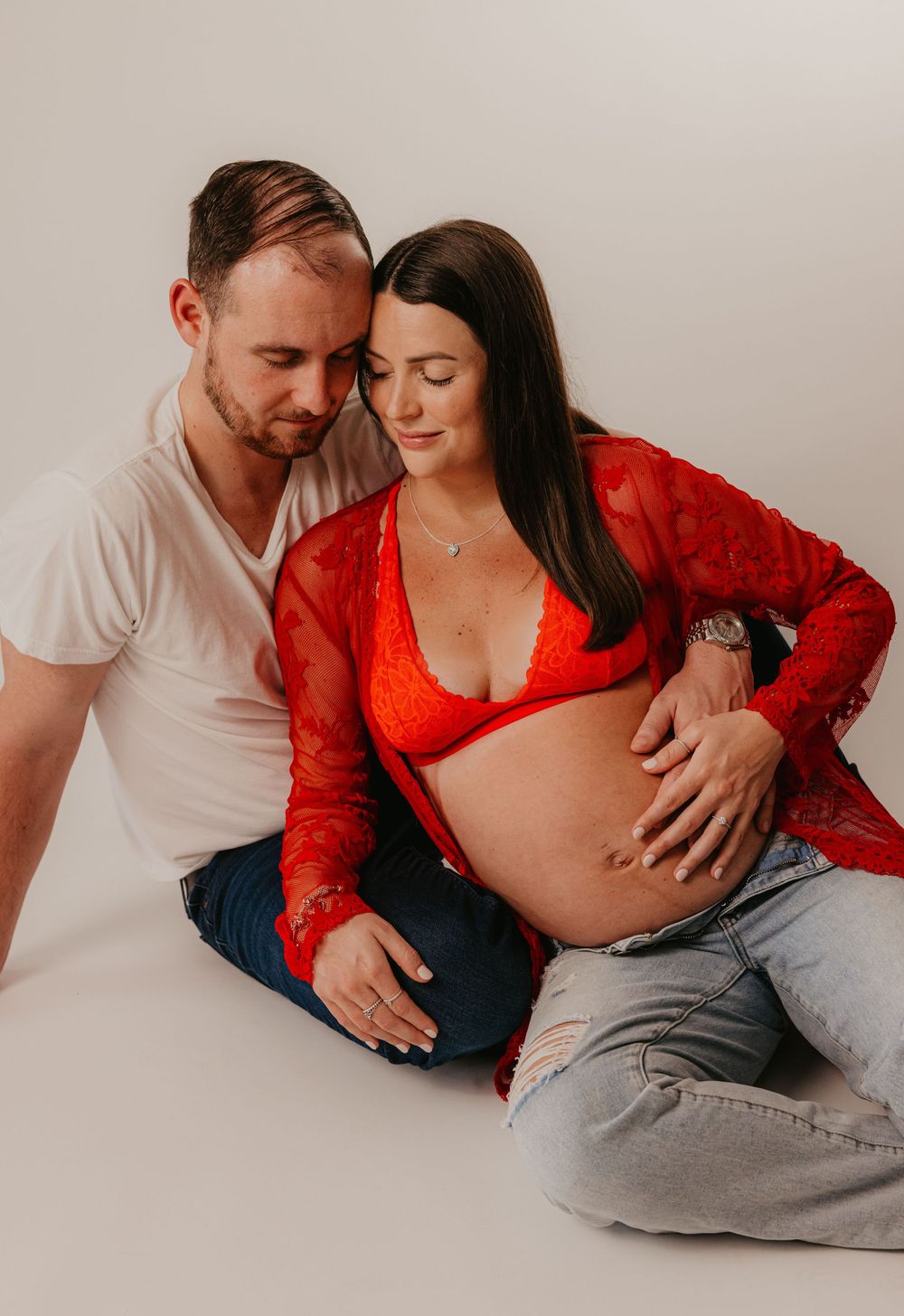Couple sitting together on a white studio floor with pregnant woman in red lace top and jeans.