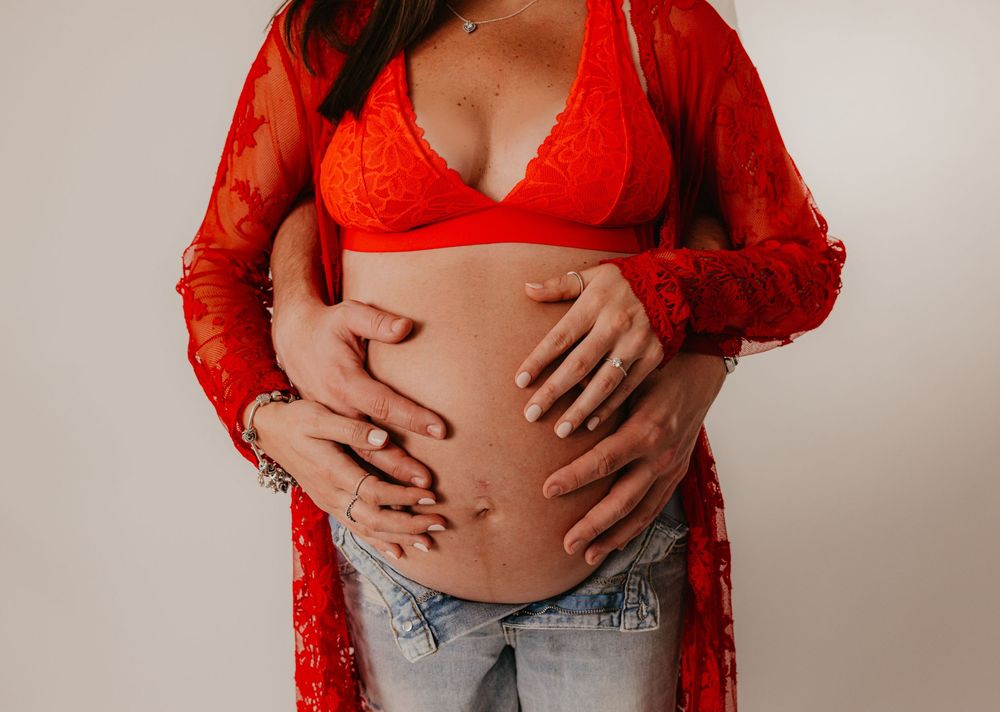 Pregnant woman in red lace bralette and kimono cradling baby bump with partner's hands on white background.
