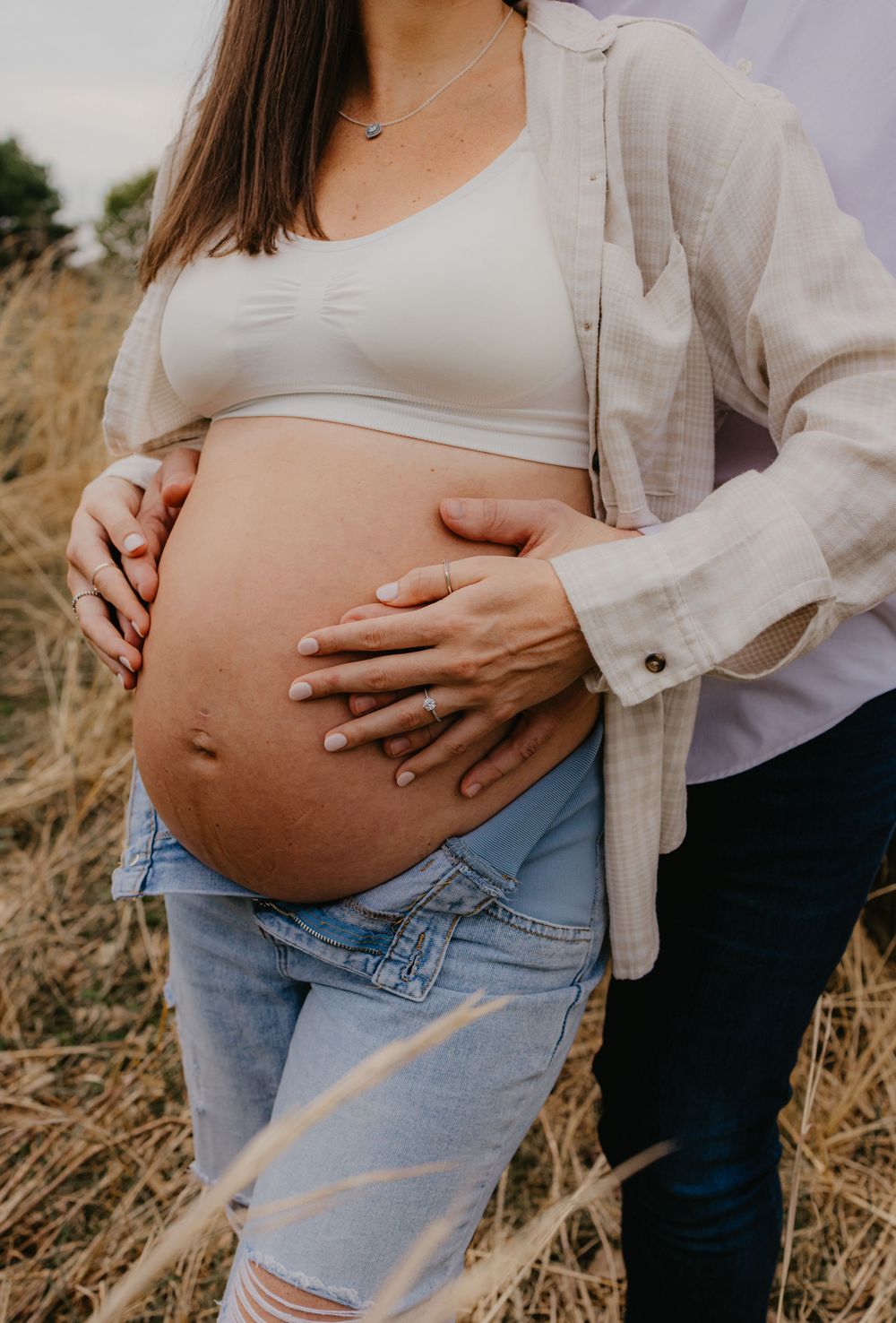 Pregnant woman in white crop top and jeans holds baby bump outdoors in golden field with partner.