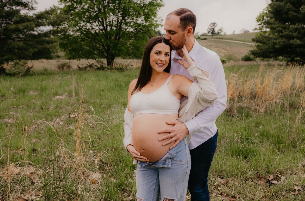 Couple poses outdoors in a field, pregnant woman in white top and jeans cradling her baby bump with partner behind her.