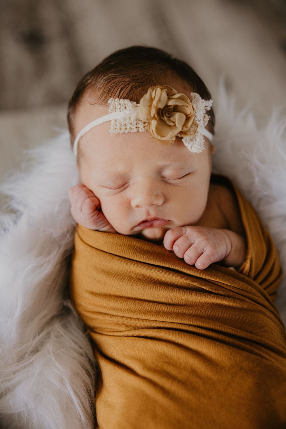 Sleeping newborn baby girl swaddled in mustard yellow wrap with white lace and gold flower headband on fluffy white blanket.