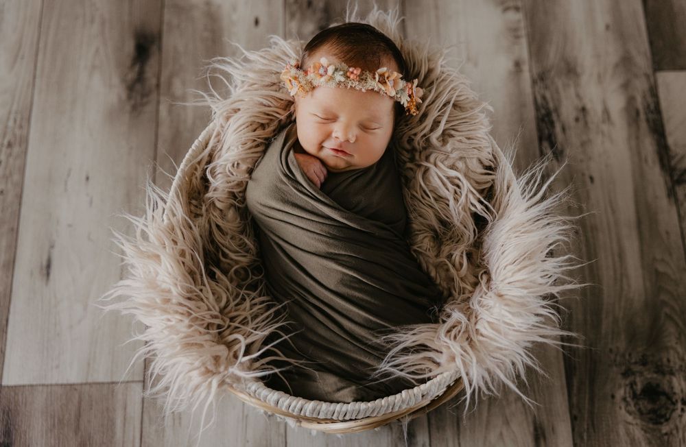 Newborn baby girl swaddled in gray wrap, resting in a fur-lined basket with a floral headband.