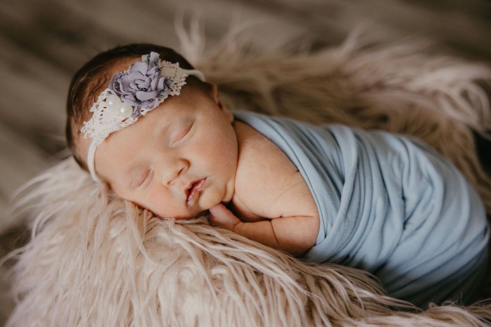 Sleeping newborn baby girl wrapped in blue swaddle with purple floral headband on fluffy fur blanket.