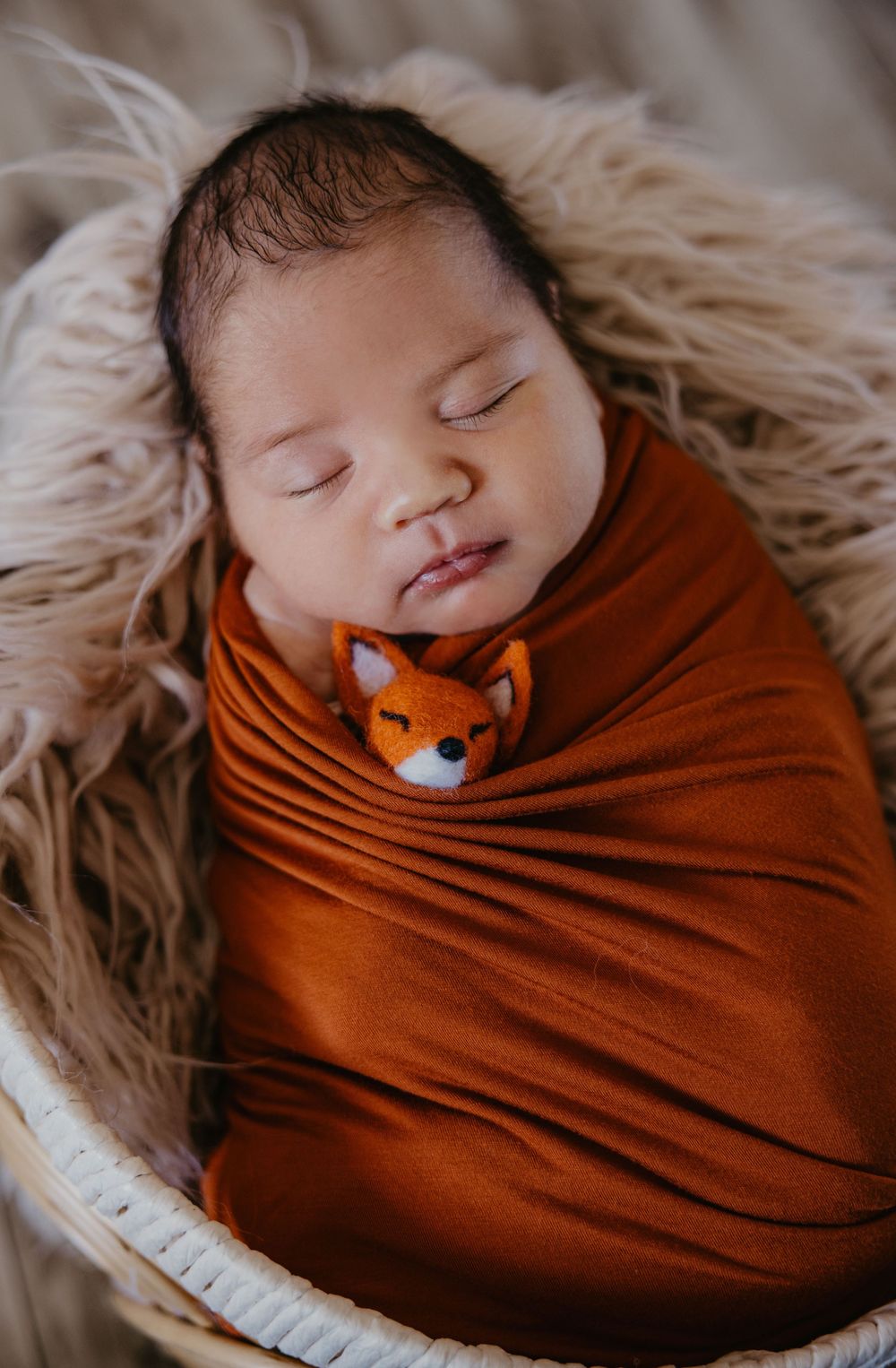 Sleeping newborn baby swaddled in rust orange wrap with a small fox toy, resting in a wicker basket.
