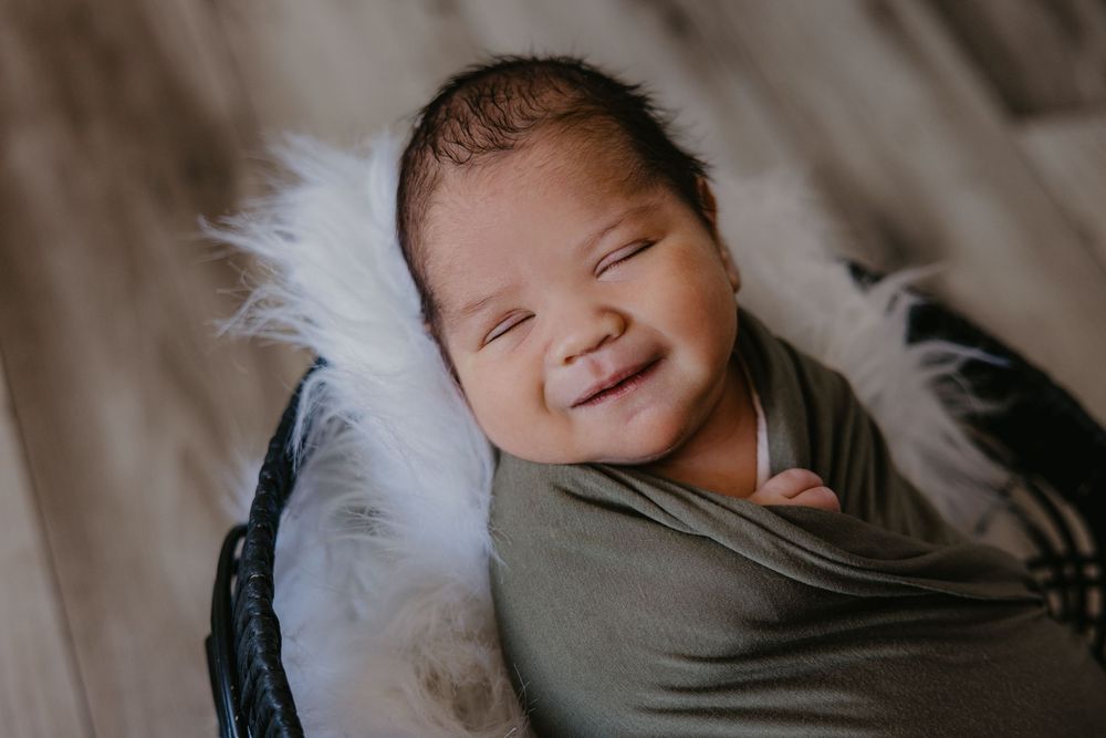 Smiling newborn baby wrapped in olive green swaddle nestled in basket with white fluffy fur, newborn photography.