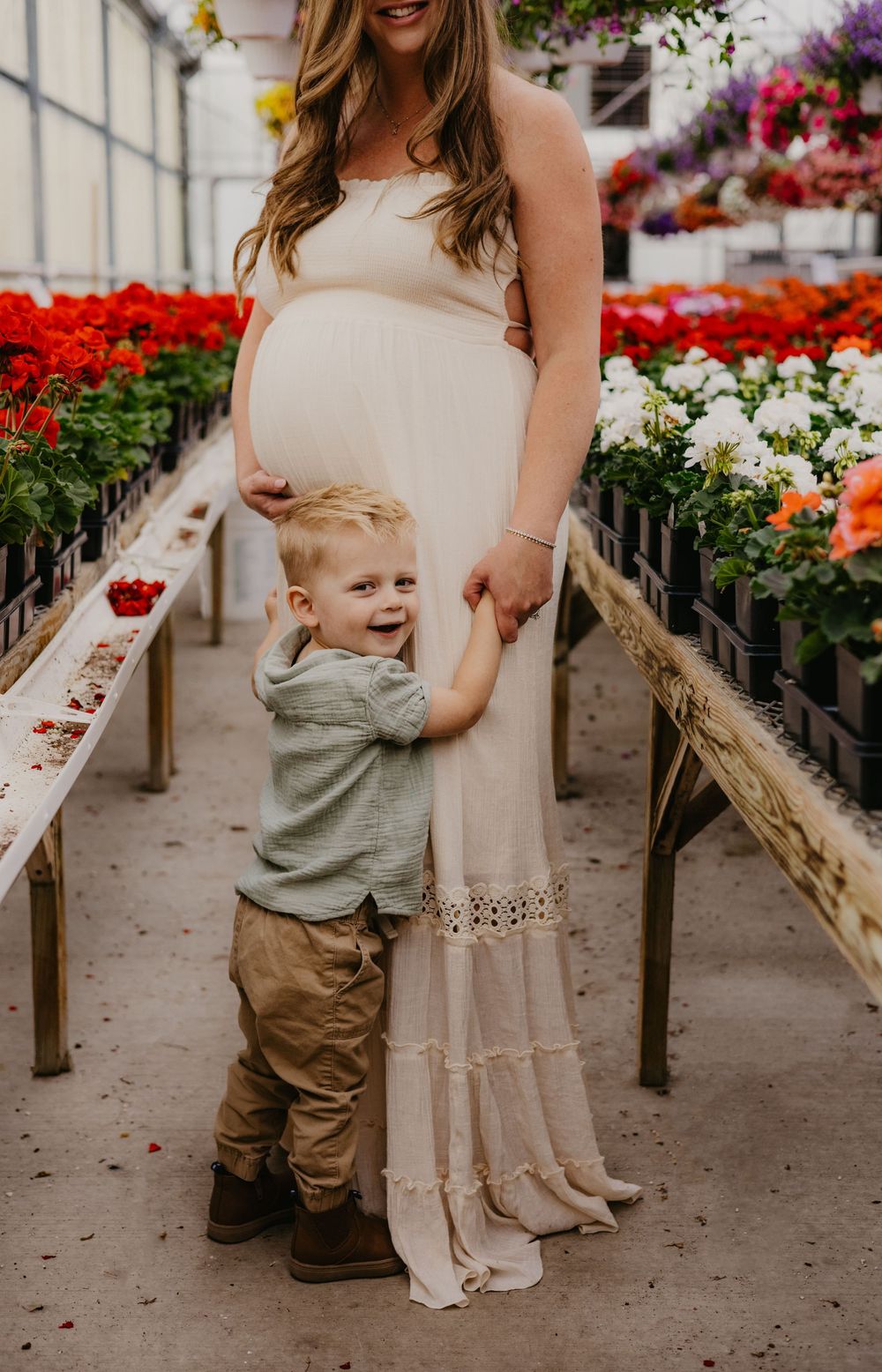 Pregnant woman in white dress with toddler boy hugging her in a colorful flower greenhouse.
