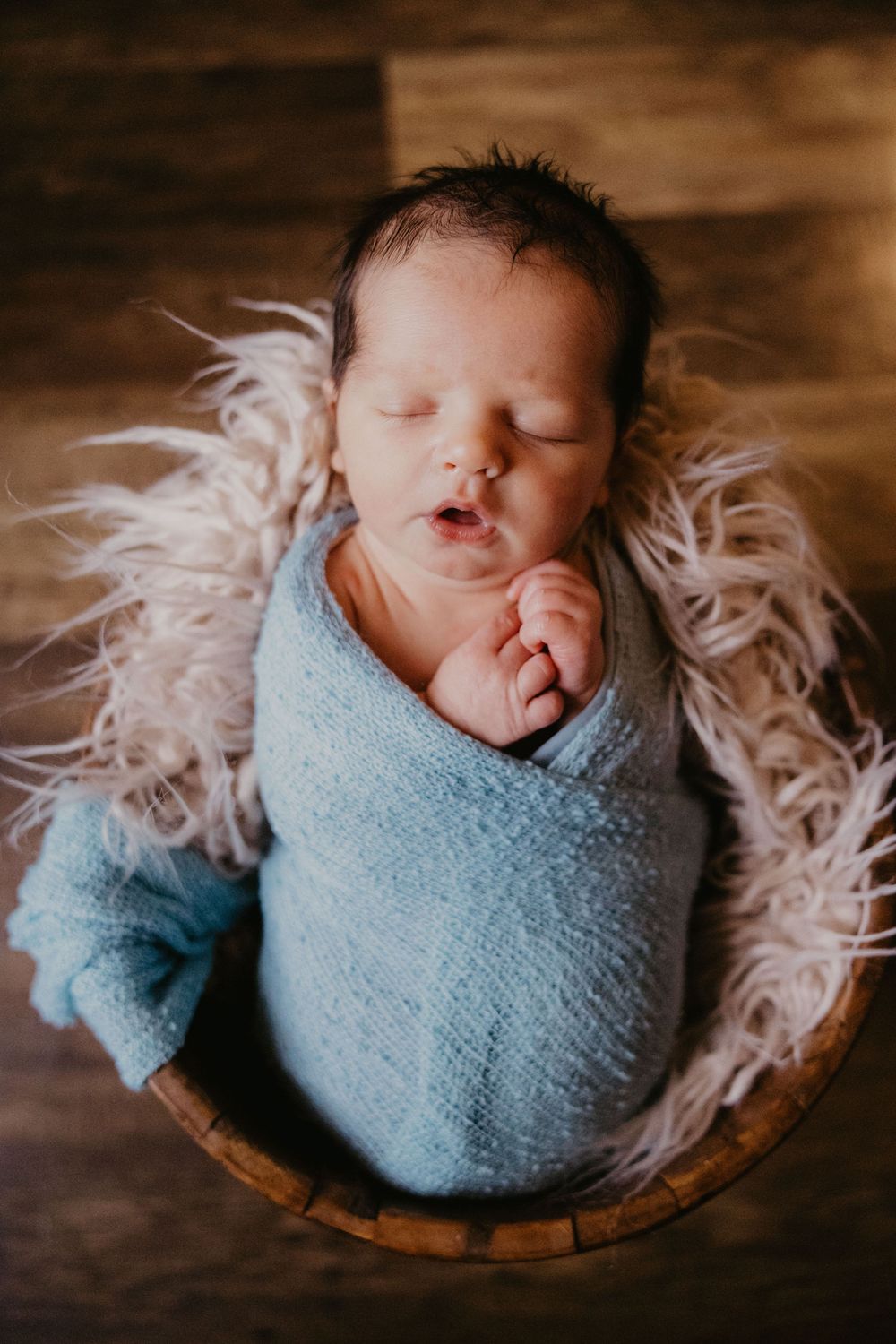 Sleeping newborn baby swaddled in blue wrap resting in a wooden basket lined with fluffy white fur.