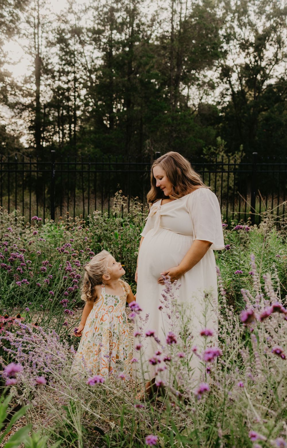 Pregnant woman in white dress stands with young daughter in floral dress among wildflowers at golden hour.