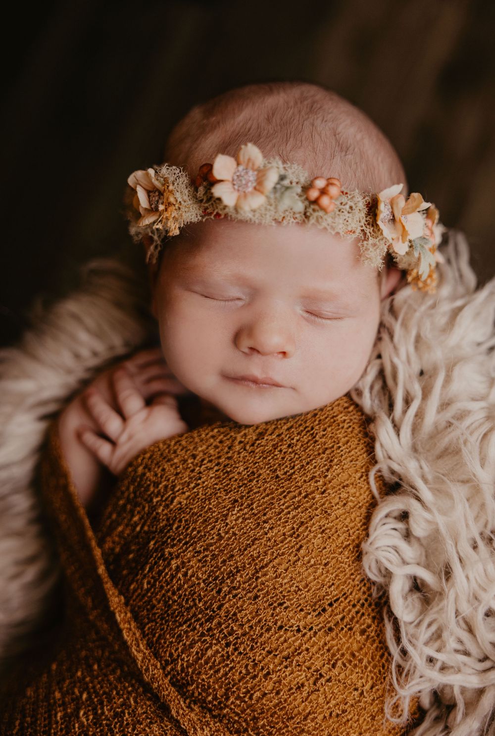 Sleeping newborn wrapped in mustard knit swaddle wearing a floral crown in a soft fur basket.
