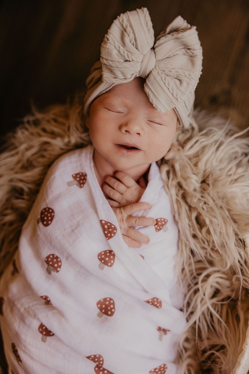 Sleeping newborn baby swaddled in white mushroom print blanket, wearing beige bow headband, resting on fluffy fur.