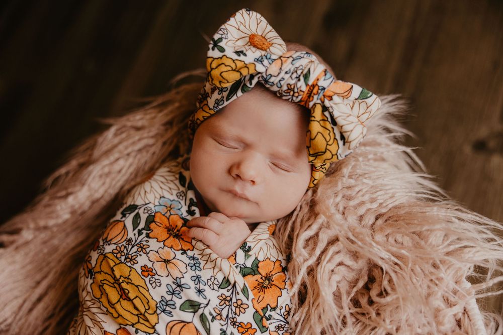 Sleeping newborn baby wrapped in floral swaddle with matching bow headband on fluffy pink blanket.