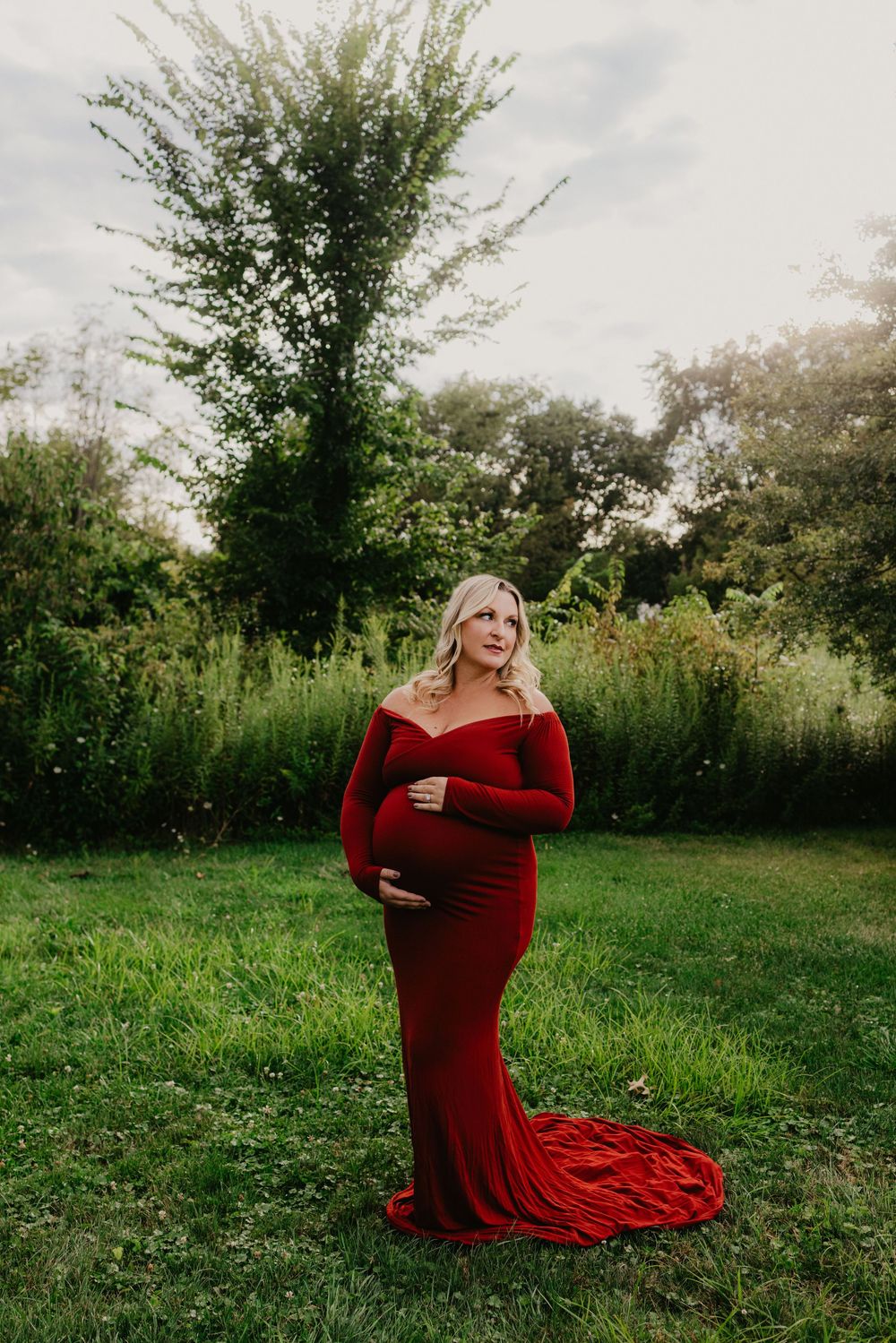 Pregnant woman in elegant red off-shoulder gown poses outdoors in lush green garden for maternity photo session.