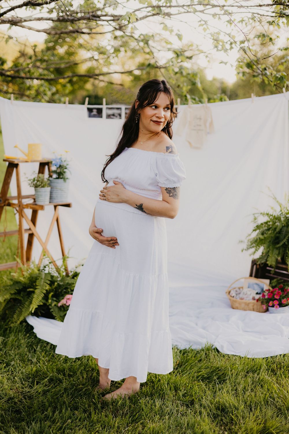 Smiling pregnant woman in a white off-shoulder dress holding her belly outdoors at a baby shower setup.