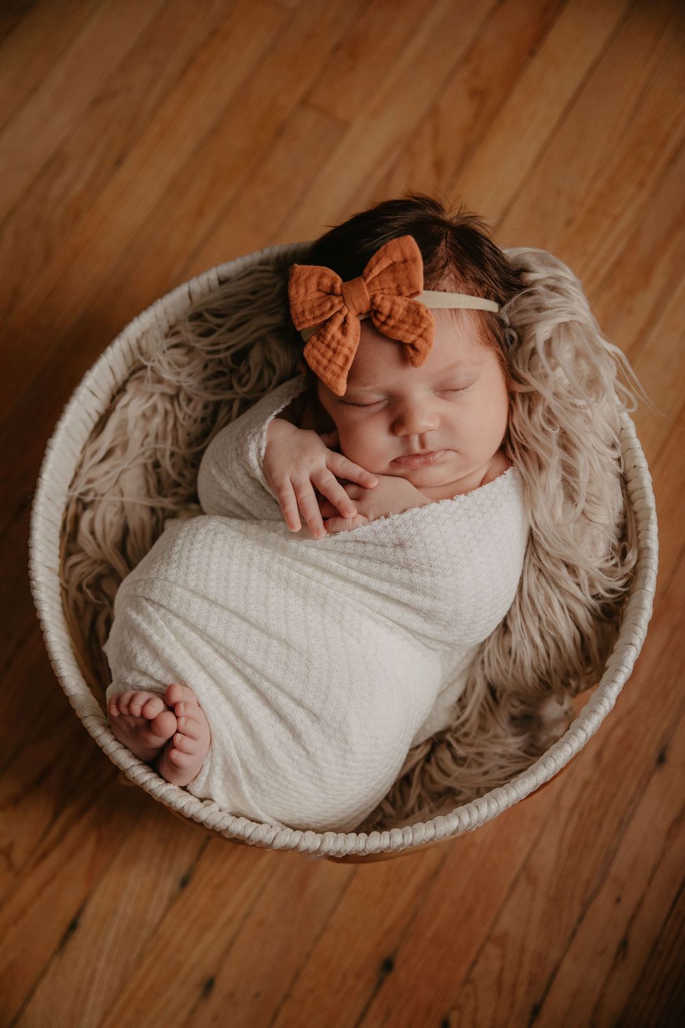 Sleeping newborn baby girl swaddled in white wrap with an orange bow headband in a round basket.