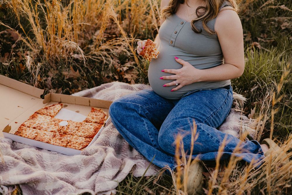 Pregnant woman sitting in a field holding her belly and a slice of pizza with an open pizza box beside her.