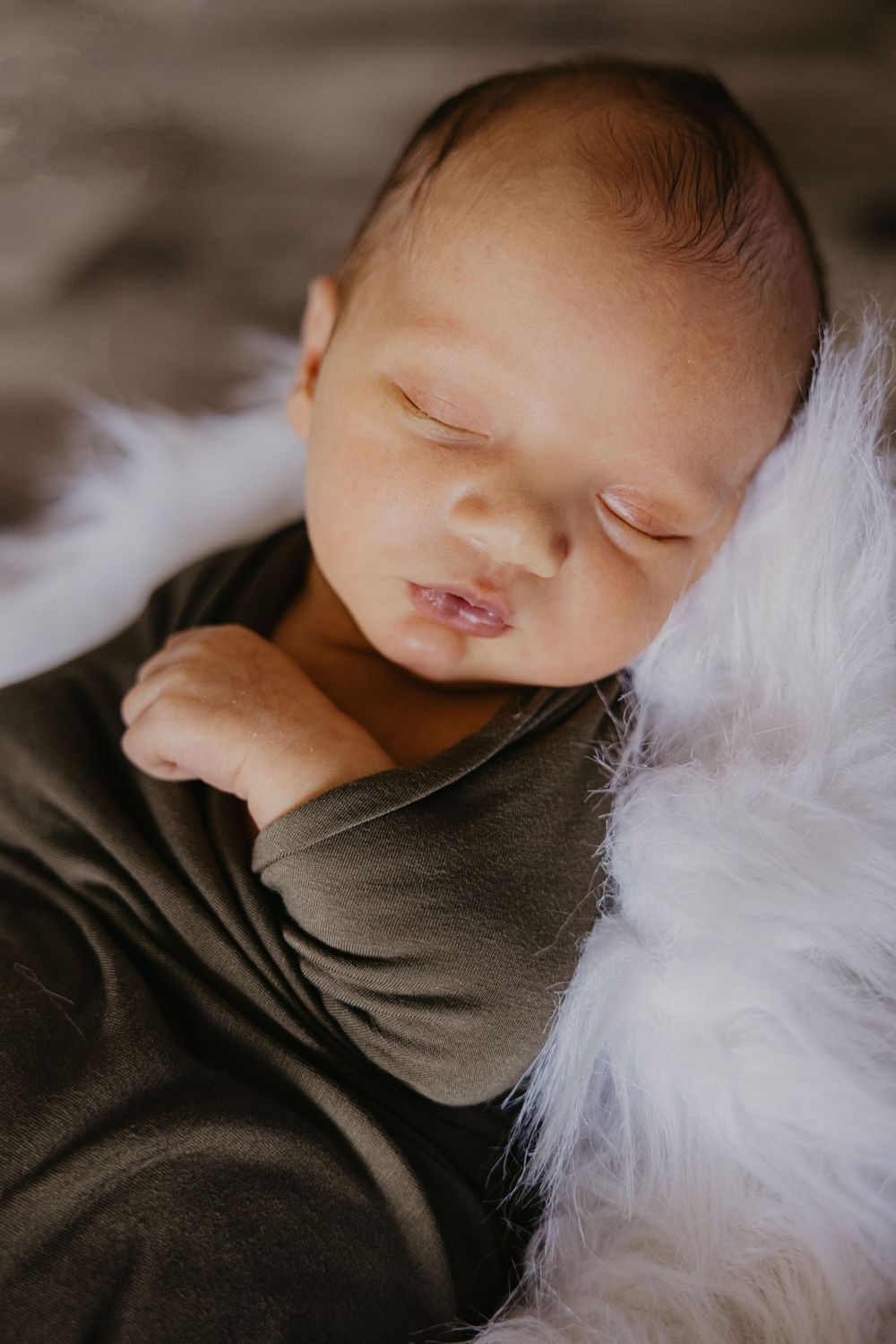 Close-up of sleeping newborn baby swaddled in gray wrap resting against white fluffy fur.