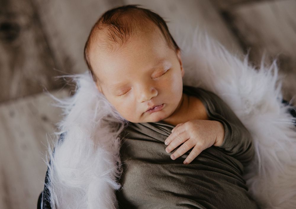 Peaceful sleeping newborn wrapped in gray swaddle nestled in white fluffy fur in a basket.
