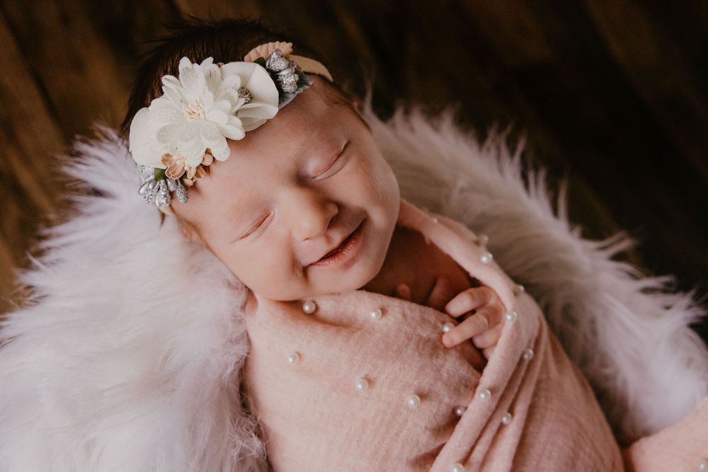 Smiling newborn baby wrapped in pink with a white floral headband nestled in fluffy fur.