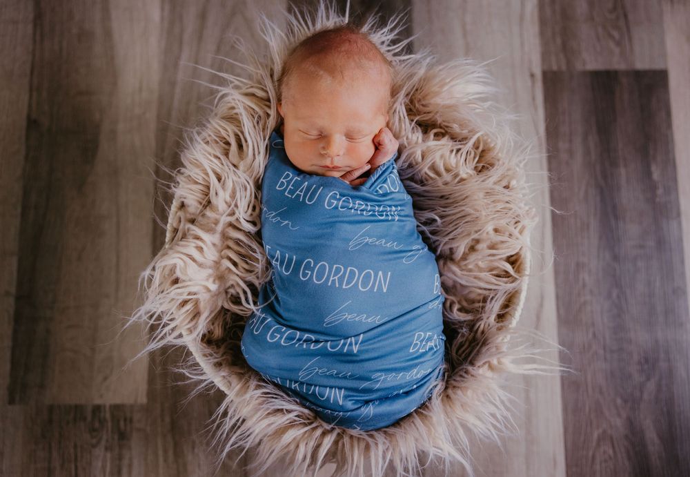 Newborn baby swaddled in blue personalized blanket reading Beau Gordon in a fluffy basket.