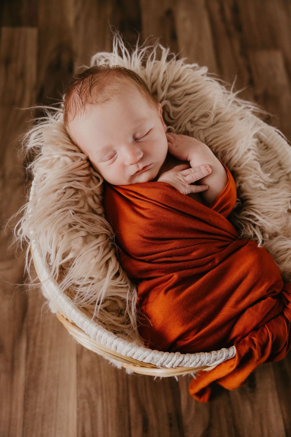 Sleeping newborn swaddled in rust orange wrap resting in a wicker basket with fluffy lining.