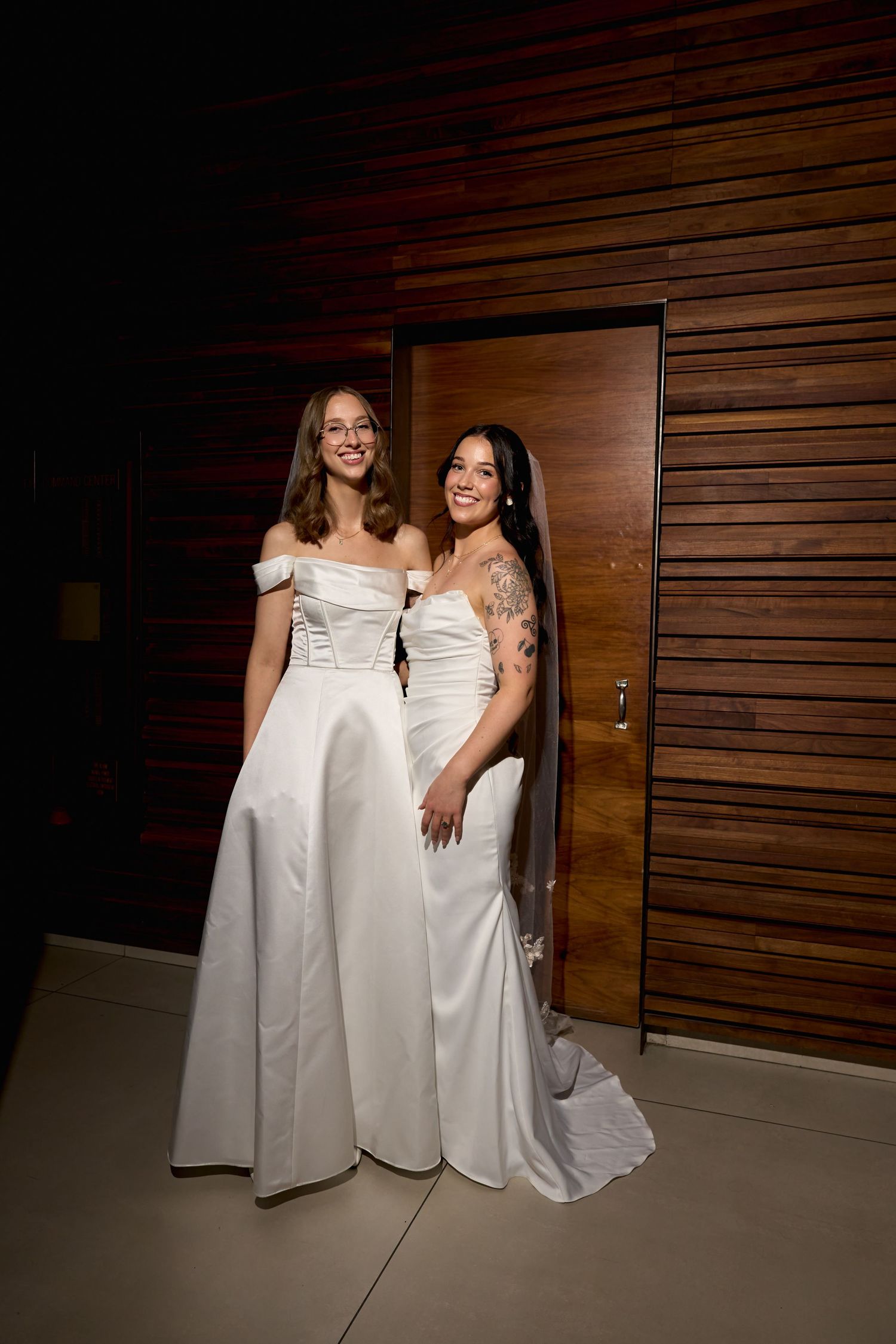 Two brides in white wedding gowns smiling together near a wooden door.
