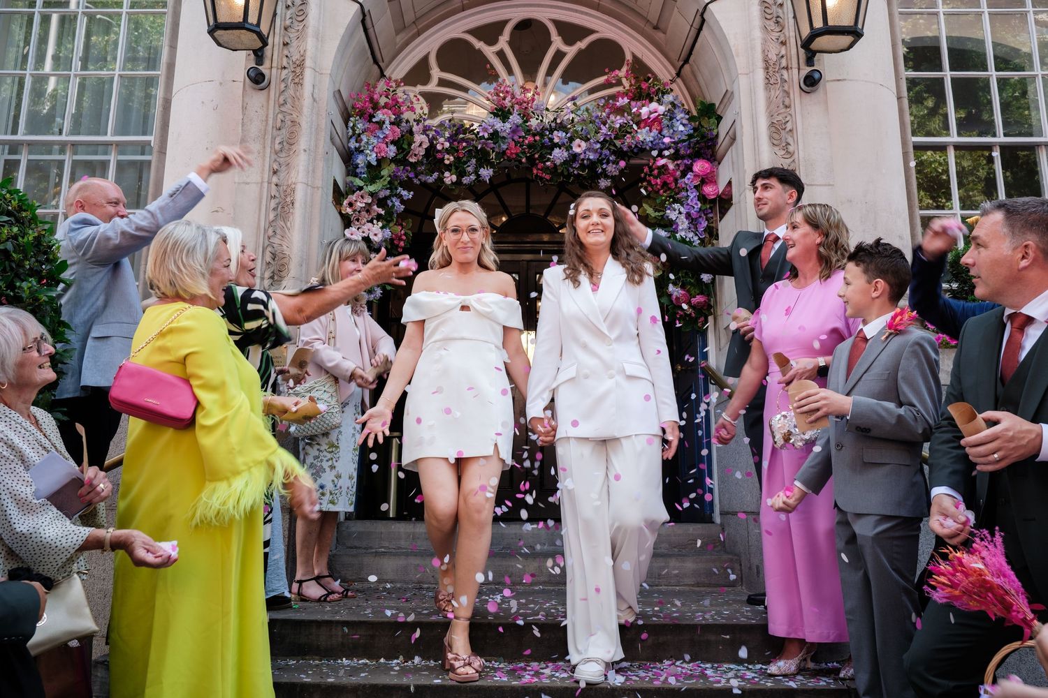 Two brides in white outfits exit a flower-adorned venue as guests celebrate with colorful confetti.