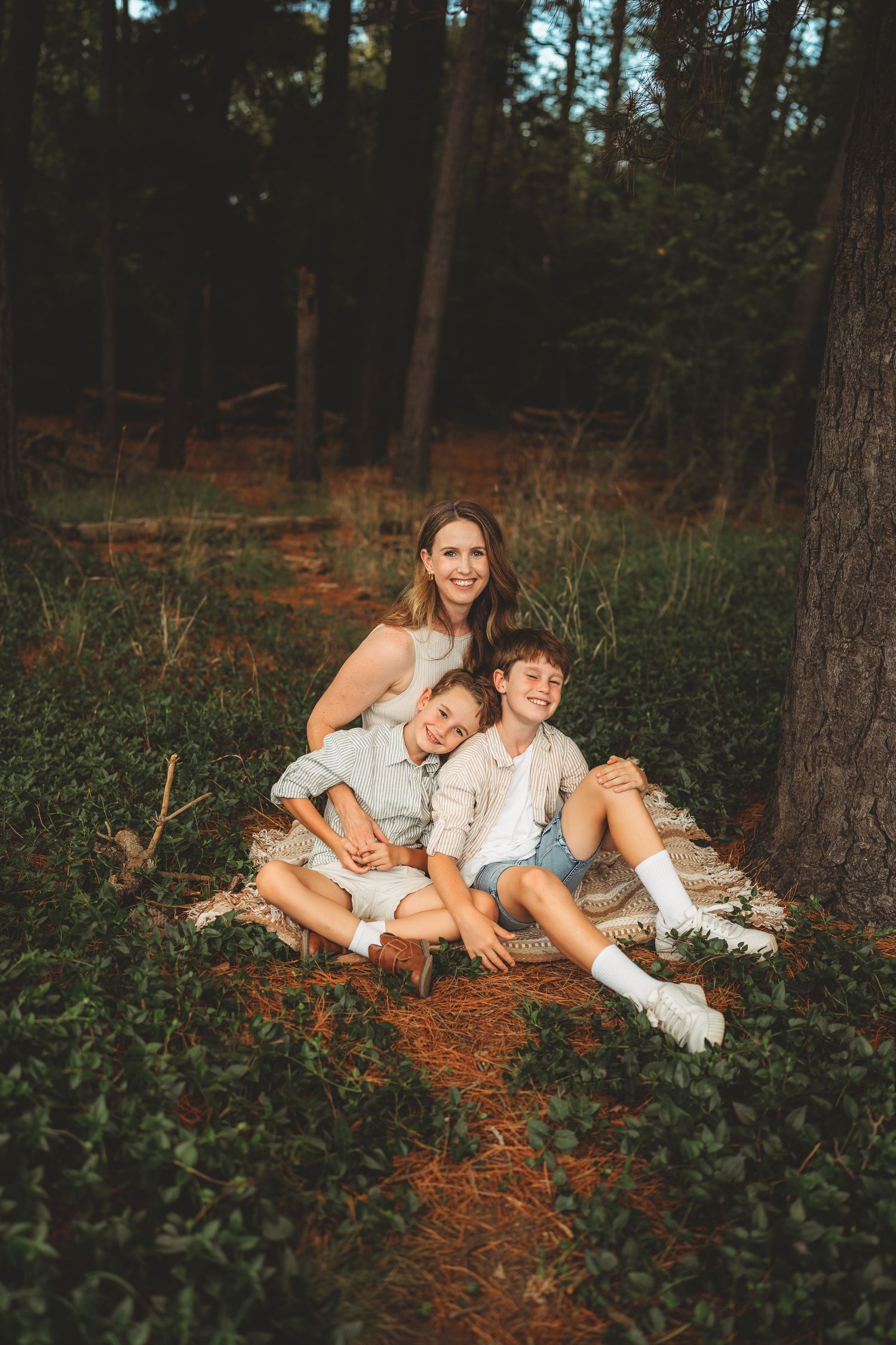 Smiling mother sitting with two young boys on blanket in pine forest during golden hour.
