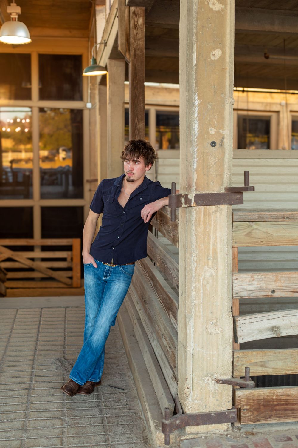 Young man in blue shirt and jeans leaning against rustic wooden post outside vintage building.