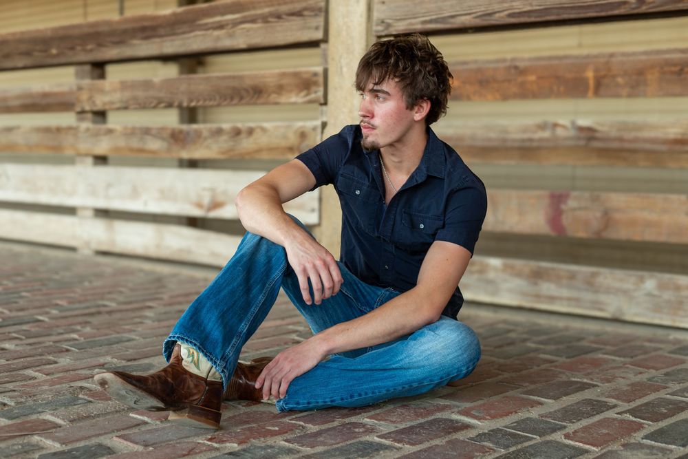Young man in blue shirt and jeans sitting relaxed on brick floor near wooden fence outdoors.