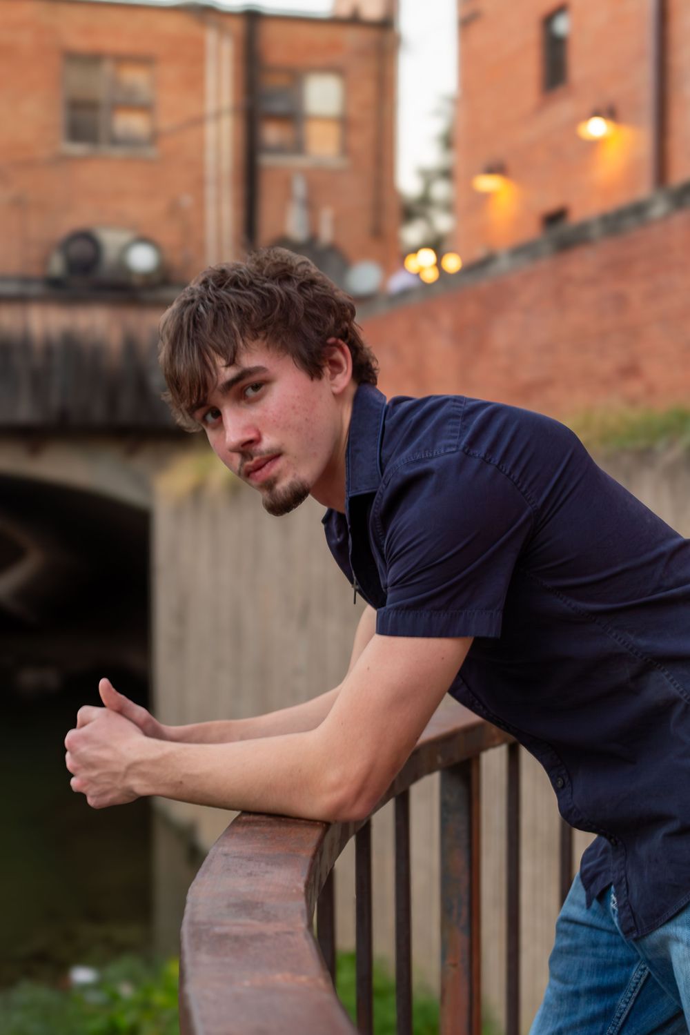 Young man in navy polo shirt leaning on urban railing near canal with brick buildings at dusk.