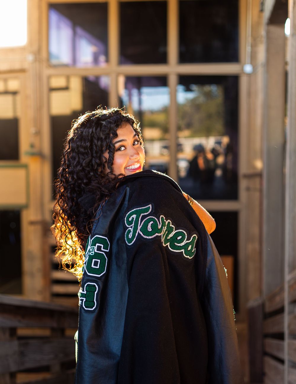 Smiling young woman with curly hair showing off a black varsity jacket with green Torres lettering outdoors.