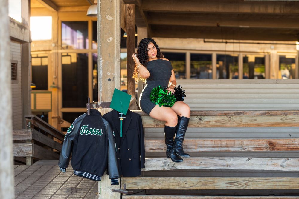 Cheerleader in black outfit and boots poses on wooden bleachers with green pom-poms and varsity jacket.