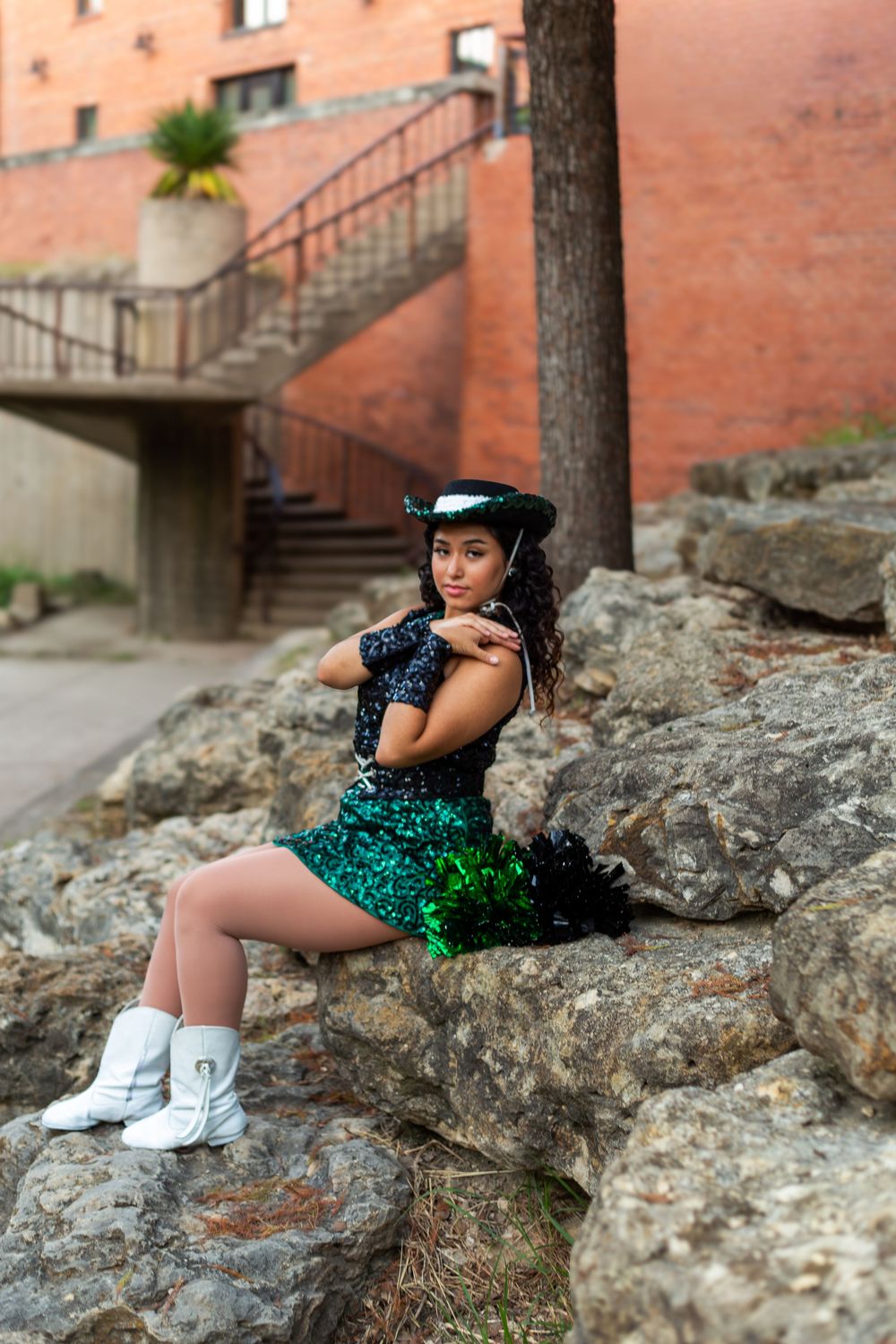 Young woman in sequined green skirt and black hat poses on rocks near brick building stairs.