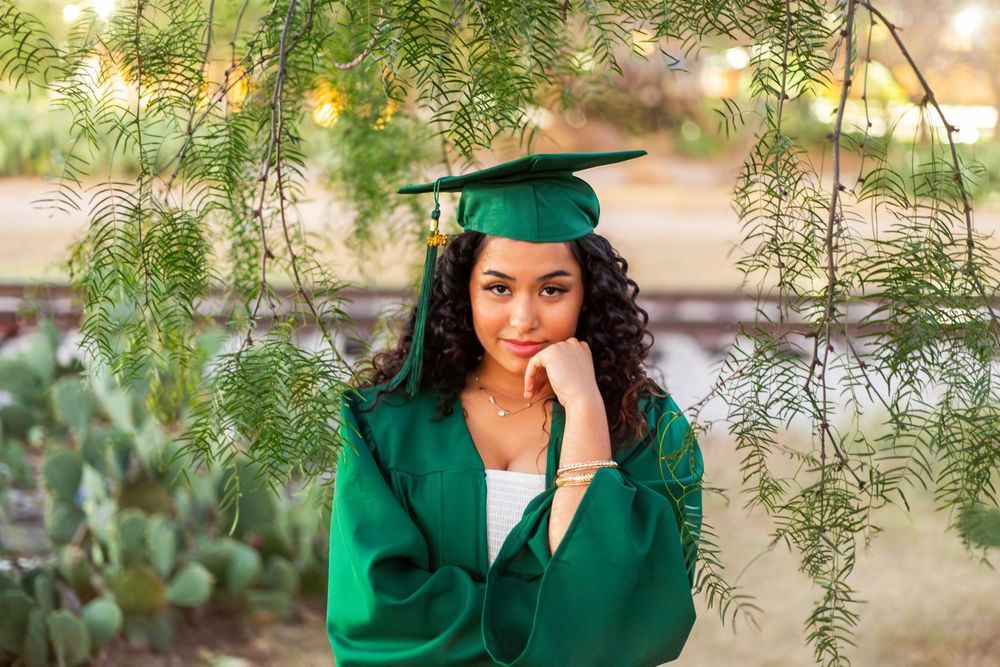 Young woman in green graduation cap and gown poses thoughtfully under hanging tree branches outdoors.
