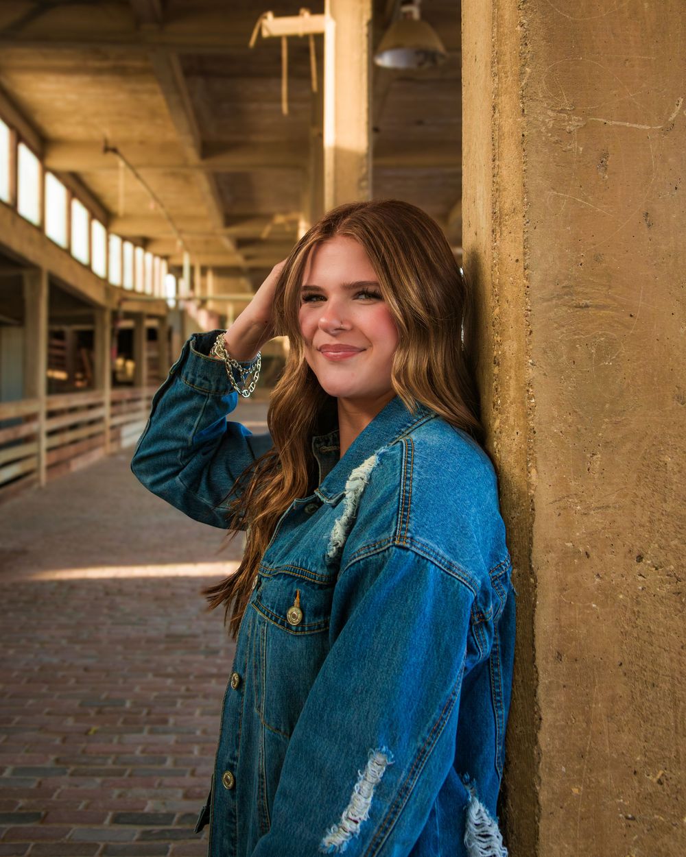 Young woman in distressed denim jacket leaning against wall smiling in urban corridor with warm lighting.