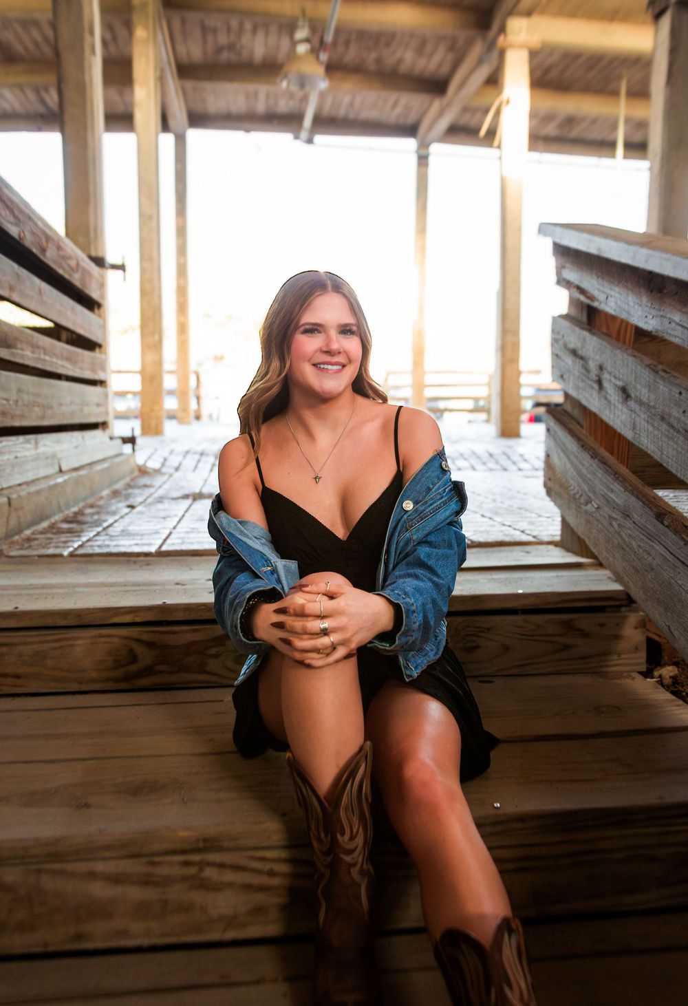 Young woman in black dress and denim jacket sitting on wooden steps wearing cowboy boots.