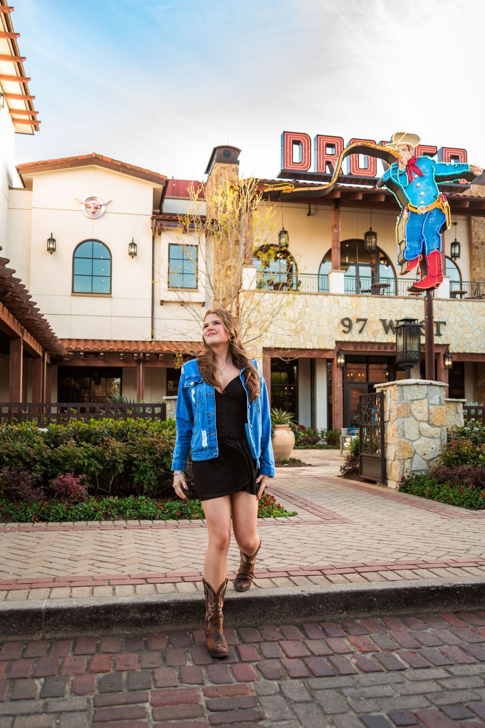Woman in blue denim jacket, black dress, and cowboy boots poses outside a Western-themed building with a neon sign.