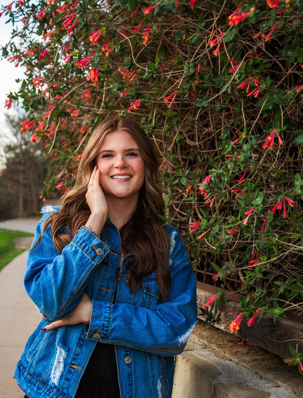 Smiling young woman in denim jacket posing by a stone wall with vibrant red flowering vines outdoors.